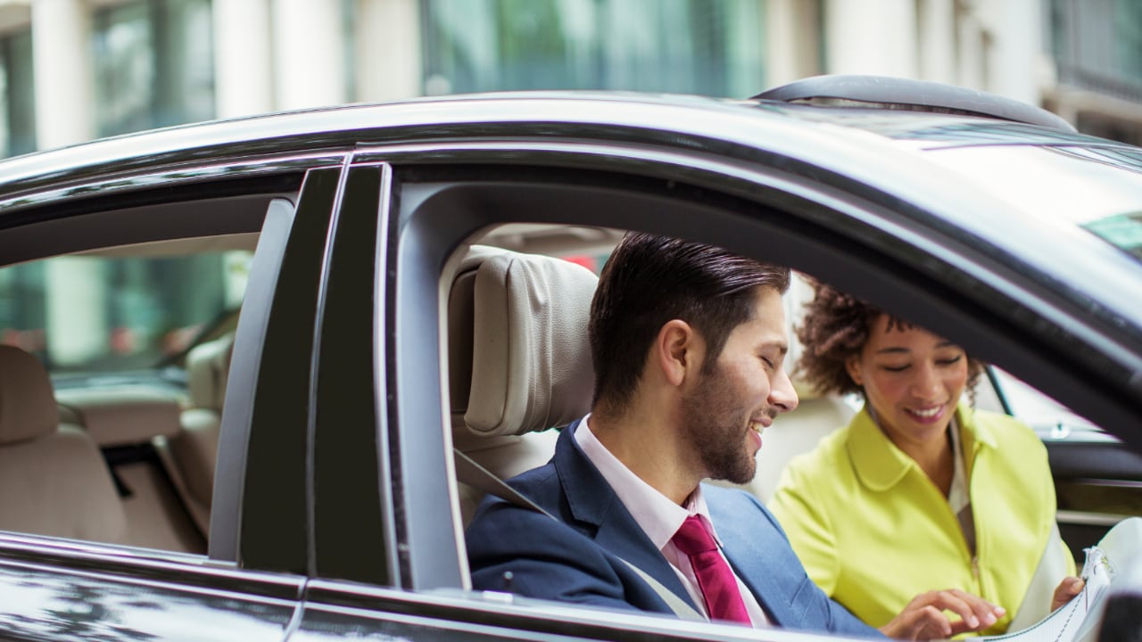Man in woman in the front of a car talking to each other.