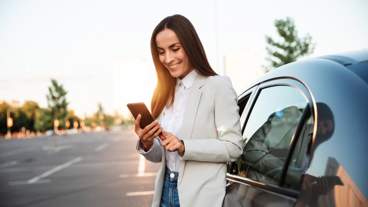 Woman smiling looking at her phone while standing outside of a car.