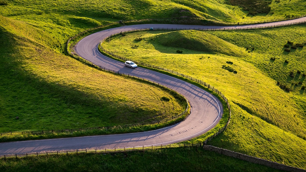 A small silver car driving round the sharp bends of a country road in the Peak District, England. Low sunlight casting shadows on the s shaped curves.