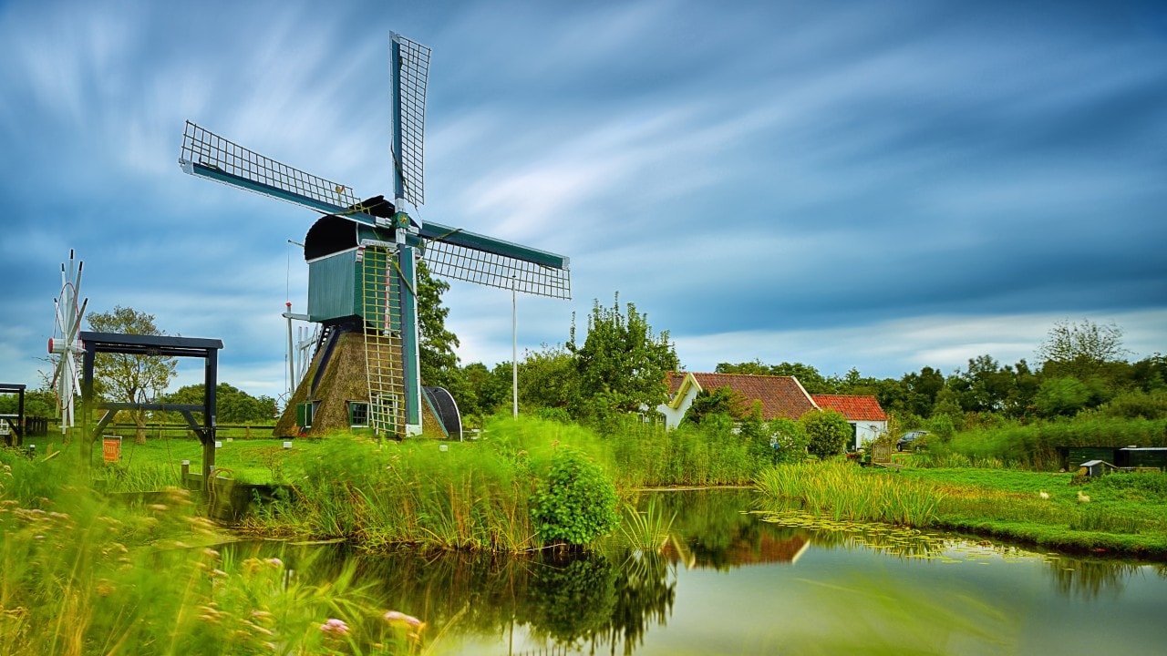 Dutch landscape with the "De Trouwe Wachter" windmill near the river in the cloudy and windy day of summer in Tienhoven, Netherlands. Long exposure with a beautiful effect in the sky. 
De Trouwe Waghter is a hollow post mill east of Tienhoven village in the Dutch province of Utrecht.
The mill was built in 1832 to replace two older mills. Until 1948 the mill has drained the polder through a paddlewheel.  The mill was restored by Dutch Nature Monuments in 1970 and 2011. The mill has rods with a length of over 20 meters that are equipped with the Old Dutch fencing with sails.