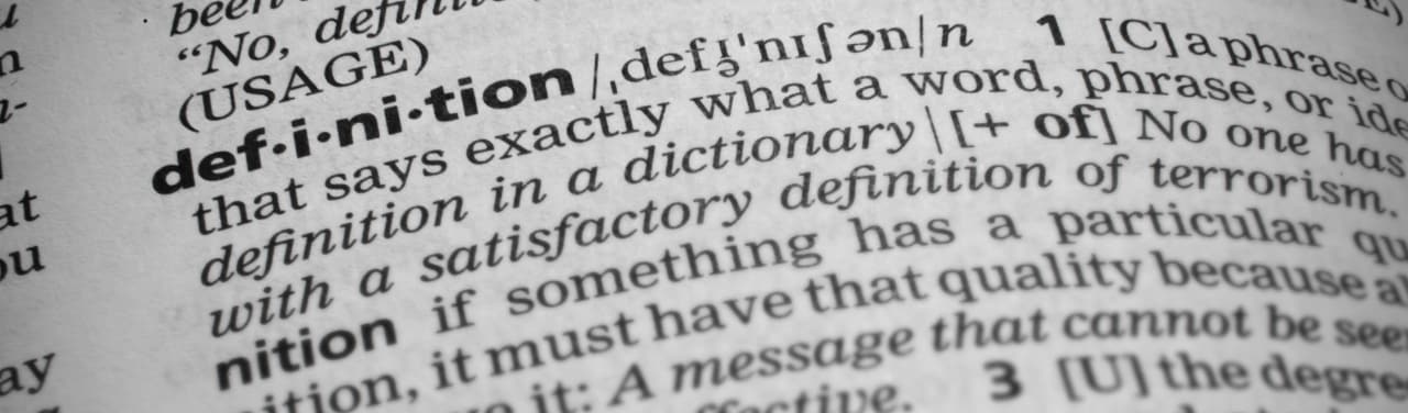 Closeup shot of the word definition in a dictionary. Page curves and has a vignetting or edge burn effect where the corners are darker. Focus is on the word definition.